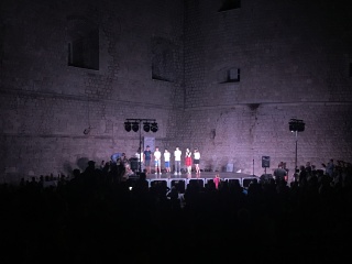 Kids on a lit stage, the fortress wall for a backdrop, crowd in silhouette.